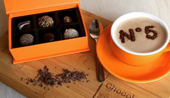 A box of chocolates and cappuccino displayed open a wooden board