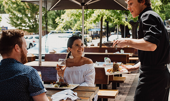 A couple are served by a waiter at a wooden outside table